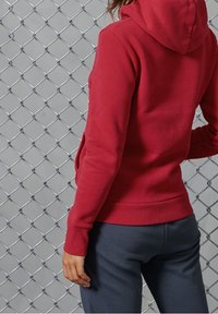 Sudadera con capucha roja de textura suave y dobladillo acanalado, combinada con pantalones de chándal grises, contra un fondo de malla de cadena plateada.