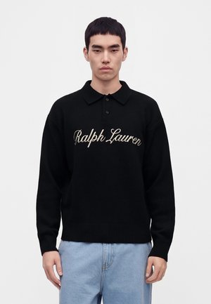Giovane uomo che indossa un maglione nero Ralph Lauren con colletto e jeans azzurri, in piedi davanti a uno sfondo chiaro e uniforme.