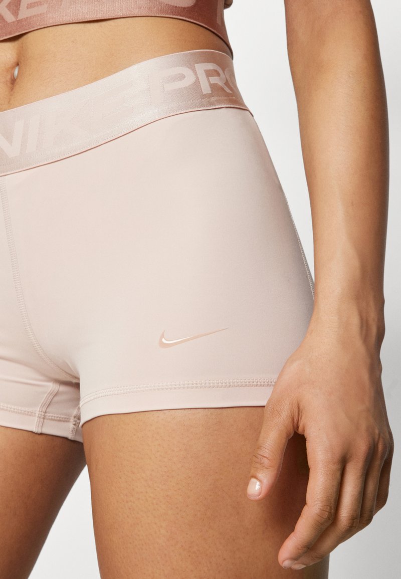Shorts Nike Pro de color rosa pálido ajustados en el muslo superior y la cintura de una persona, mostrando el logo de Nike y una mano descansando al lado de la pierna.