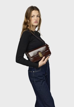 Jonge vrouw in een zwarte top met lange mouwen en donkere spijkerbroek die een donkerbruine leren schoudertas met gouden sluiting vasthoudt.