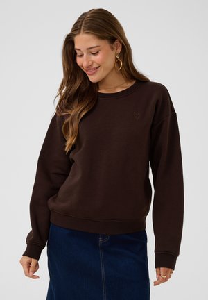 Femme aux longs cheveux bruns portant un pull marron foncé et une jupe en denim bleue, souriante et regardant vers le bas, les mains détendues le long du corps.