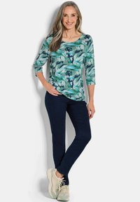 Bloemrijk, teal bovenstuk met driekwart mouwen, gecombineerd met donkerblauwe broek en lichte sneakers, met een ronde halslijn.