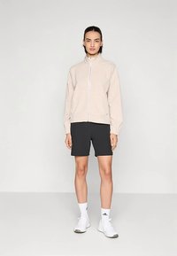 Svetlosmeđa jakna s uzorkom i patentnim zatvaračem, uparena s crnim kratkim hlačama. Bijele sportske tenisice zaokružuju izgled. Jednostavan dizajn i lagani materijal.