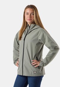 Junge Frau mit langen Haaren trägt eine hellgraue Kapuzenjacke mit Frontreißverschluss und Reißverschlusstaschen an den Seiten, kombiniert mit blauen Jeans.