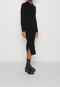 Robe noire côtelée à manches longues, col montant et fentes latérales. Associée à des bottes noires à talons épais. Texture lisse et design ajusté.