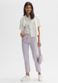 Giacca di jeans bianca tagliata, top bianco semplice, pantaloni lavanda con cordoncino e sneakers bianche. Tiene una piccola borsa intrecciata lavanda.