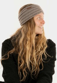 Fascia grigia lavorata a maglia con texture a coste orizzontali e un'etichetta in pelle marrone, indossata da una persona con capelli lunghi e mossi, su uno sfondo bianco.