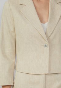 Blazer beige en tissu texturé, avec un col à revers, fermeture à un bouton et manches raccourcies.