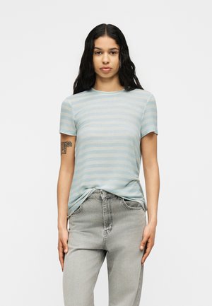 Giovane con lunghi capelli neri che indossa una maglietta a righe azzurro chiaro e jeans grigi, in piedi con una mano in tasca davanti a uno sfondo semplice.