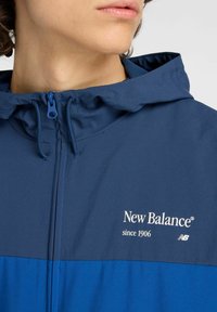 Veste imperméable bleu marine avec capuche ajustable, fermeture éclair et logo "New Balance" blanc. Présente un design de panneaux contrastants bleus.