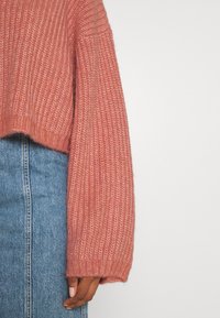 Stickad, kort tröja i dammig rosafärg. Har långa, lösa ärmar och en ribbad textur. Matchas med högmidjade blå denim.