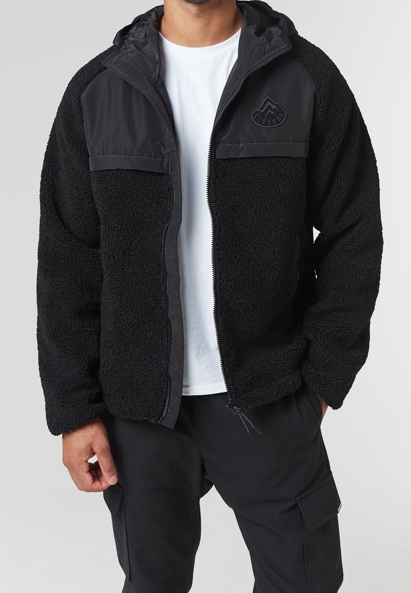 Homme portant une veste polaire noire avec fermeture éclair, un pantalon cargo noir et un t-shirt blanc, debout les mains détendues le long du corps.