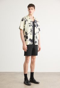 Camicia a maniche corte di un giallo chiaro con un motivo floreale nero, abbinata a pantaloni corti neri, calze nere e scarpe nere. Modello in piedi.