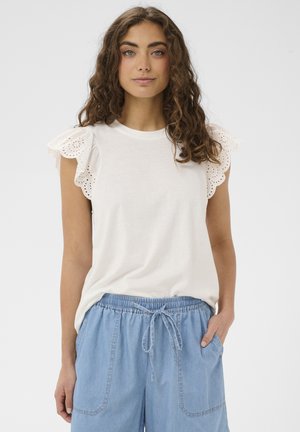 Femeie cu păr creț purtând un top alb fără mâneci cu mâneci dantelate volanate și pantaloni din denim albastru deschis cu șnur ajustabil.