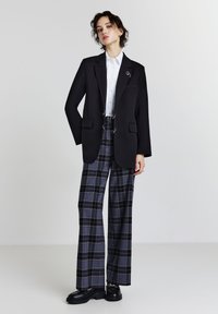 Blazer nero su misura con due tasche frontali, abbinato a pantaloni a quadri larghi in grigio e blu navy. Camicia bianca con colletto sotto, scarpe nere.