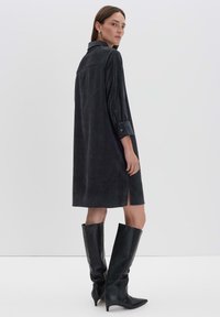 Vrouw in een losse, donkergrijze corduroy jurk met 3/4 mouwen en zwarte leren laarzen met een gemiddelde hak tot aan de knie, staande tegen een effen achtergrond.