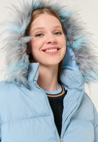 Giubbotto puffer azzurro chiaro con cappuccio removibile, caratterizzato da un bordo in pelliccia sintetica grigia e azzurra chiara. Zip visibile e colletto a coste.
