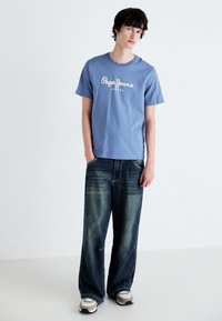 Šviesiai mėlyna medvilninė marškinė su "Pepe Jeans London" spaudiniu, derinama su tamsiai mėlynomis plačiomis džinsinėmis kelnėmis ir smėlio bei juodos spalvos sportiniais bateliais.