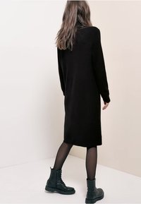 Schwarzes Strickkleid mit langen Ärmeln, knielanges Design, glatte Textur. Kombiniert mit schwarzen Kampfstiefeln und schwarzen, transparenten Strumpfhosen.