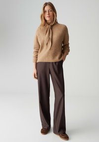 Pull beige en tricot avec un lien au col, associé à un pantalon large marron foncé et des chaussures marron, debout devant un fond clair.