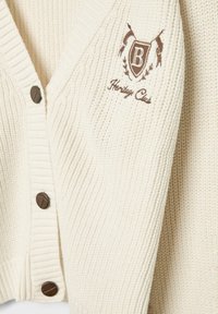 Cardigan côtelé de couleur crème avec un col en V, des boutons marron et un blason brodé comportant le texte "Heritage Club".