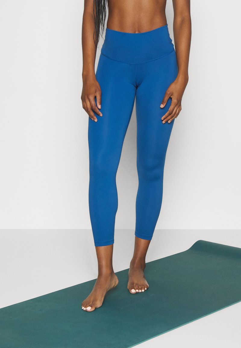 Blå leggings av elastiskt, slätt tyg med hög midja och ankellängd. Modellen står barfota på en grön yogamatta.