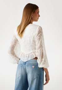 Witte geborduurde blouse met doorzichtige lange mouwen en een peplumzoom, gecombineerd met lichtblauwe high-waisted jeans met een driehoekig logo-opdruk.