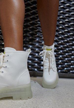 Personne portant des bottines blanches à lacets avec des semelles translucides, debout sur un sol gris clair devant un fond métallique texturé.