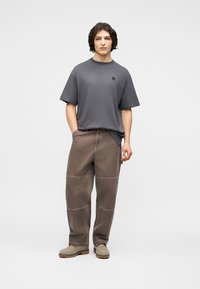 Νέος άνδρας στέκεται με τα χέρια στις τσέπες, φορώντας ένα άνετο γκρι μπλουζάκι, καφέ παντελόνια εργασίας και μπεζ μποτάκια σε λευκό φόντο.