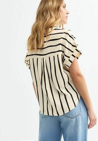 Chemise beige à manches courtes avec des rayures verticales noires, dotée d'une pli au dos et d'un ourlet incurvé, associée à un jean en denim bleu clair.