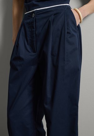 Donna che indossa pantaloni blu navy a vita alta con pieghe e tasche, una mano in tasca, indossa un braccialetto color crema al polso.