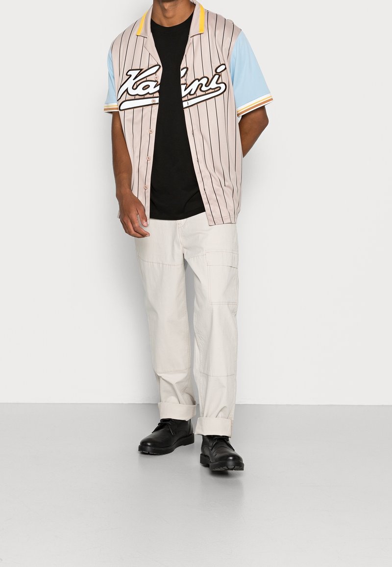 Maglietta stile baseball beige con strisce nere, con un grande logo bianco, maniche corte blu e colletto giallo. Abbinata a pantaloni cargo chiari e scarpe nere.