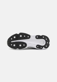 Semelle de chaussure avec un fond en caoutchouc noir présentant un motif texturé et une semelle intermédiaire blanche. Des tampons de traction circulaires et des détails de marque sont visibles.