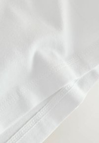 Valkoinen puuvillainen T-paita, jossa on sileä pinta, hienovaraiset rypyt ja tasaisesti ommellut reunat, korostaen yksinkertaisuutta ja hillittyjä suunnitteluelementtejä.