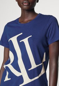 Osoba v modrém tričku s velkými bílými překrývajícími se písmeny "R" a "L" vytisknutými na přední straně, viditelné od ramen až k pasu.