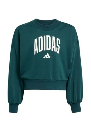 Giovane che cammina con felpa e pantaloni Adidas verde scuro e sneakers bianche su sfondo bianco.