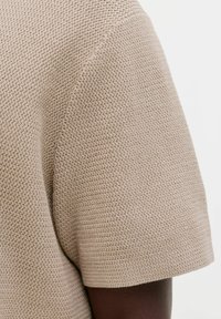 Gros plan sur un tricot beige texturé à manches courtes porté sur un bras à la peau foncée, sur un fond clair uni.
