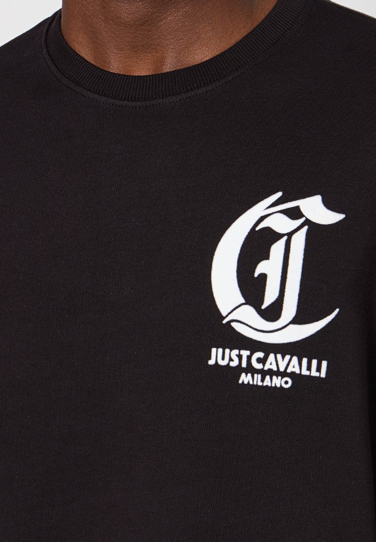 Czarna bawełniana koszulka z białym logo oraz stylizowanymi literami "J" i "C" oraz napisem "JUST CAVALLI MILANO" poniżej. Prosty, okrągły dekolt.