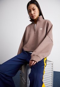 Sweatshirt castanho, cortado e com fecho de zíper a um quarto, feito de um tecido suave; apresenta o logótipo. Combinado com calças azul-marinho, realçadas com riscas amarelas.