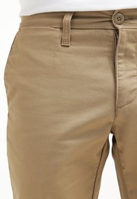 Pantaloni in cotone color khaki con una texture liscia, dotati di chiusura con bottone e passanti per cintura sulla vita, mostrati dalla vita in giù.