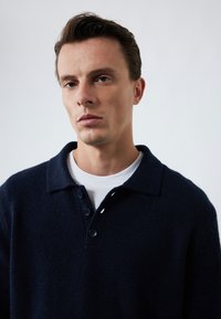 Mørkeblå strikket pullover med krave, forsynet med fire knapper og et struktureret stof; båret over en hvid T-shirt med rund hals.