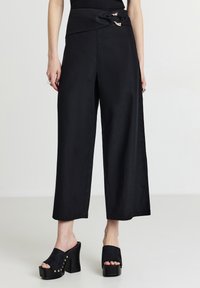 Donna con pantaloni neri a gamba larga e taglio corto, con una chiusura decorativa, e sandali mule con tacco platform neri, in piedi su uno sfondo semplice.