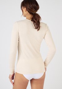 Haut à manches longues en maille côtelée de couleur beige clair, doté d'un col rond et d'une coupe ajustée. Le dos est lisse avec un ourlet légèrement plus long.