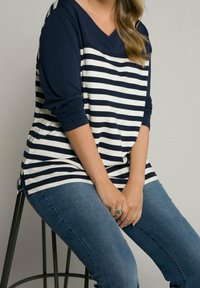 Donna seduta su uno sgabello, indossa una camicia a righe navy e bianca a maniche lunghe e jeans blu, con le mani unite in grembo.