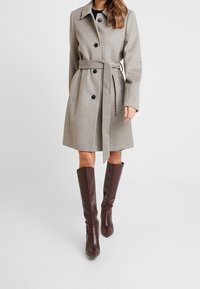 Abrigo de mezcla de lana gris claro con cintura ceñida, que presenta botones negros y cuello con muesca. Combinado con botas de rodilla en burdeos oscuro.