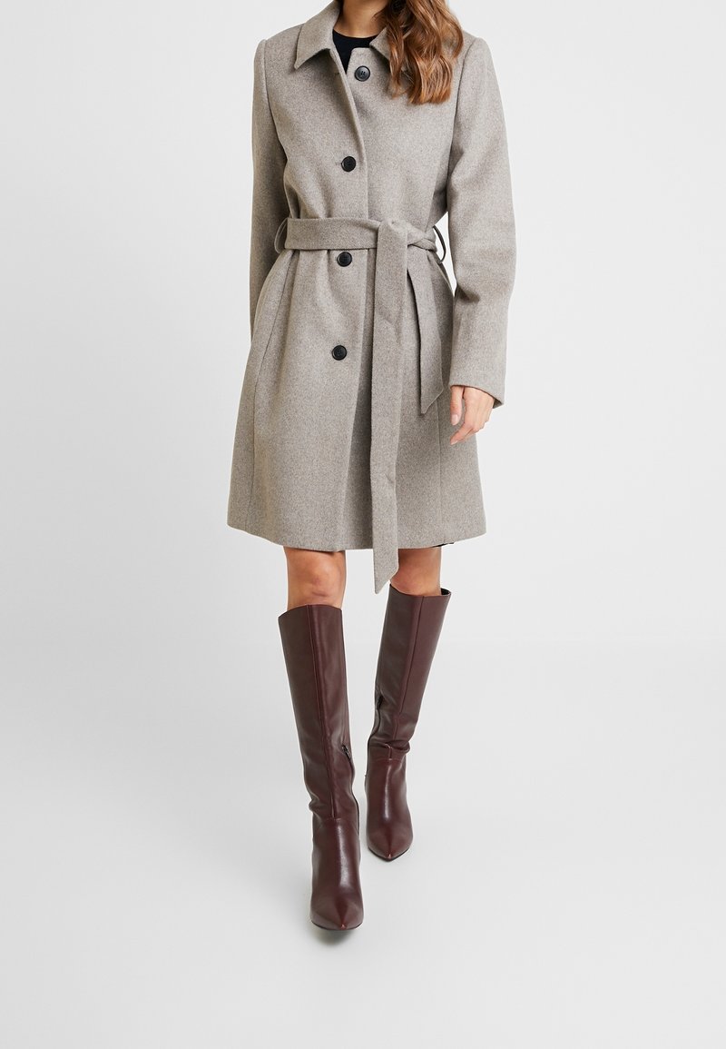 Abrigo de mezcla de lana gris claro con cintura ceñida, que presenta botones negros y cuello con muesca. Combinado con botas de rodilla en burdeos oscuro.