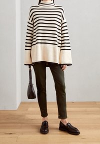 Sweter we wzory w kremowo-czarnym kolorze, o oversize'owym kroju z ściągaczami. Połączony z ciemnozielonymi spodniami i czarnymi loafersami, trzymając małą torbę.