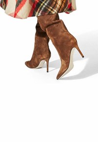 Botas de tacón de aguja altas de ante marrón con punteras puntiagudas y un tacón delgado, combinadas con una falda de cuadros multicolor que presenta tonos rojos y amarillos.
