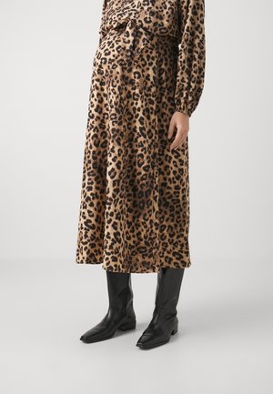 Mujer con falda midi de estampado de leopardo, top a juego y botas negras de cuero hasta la rodilla sobre un fondo liso.