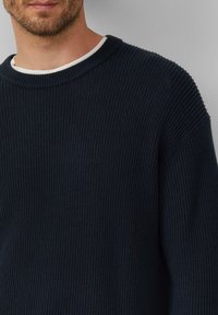 Mornarska rebrasta pletena jopica z okroglim izrezom in spuščeno ramo, z belim majico pod jakno vidno na ovratniku.